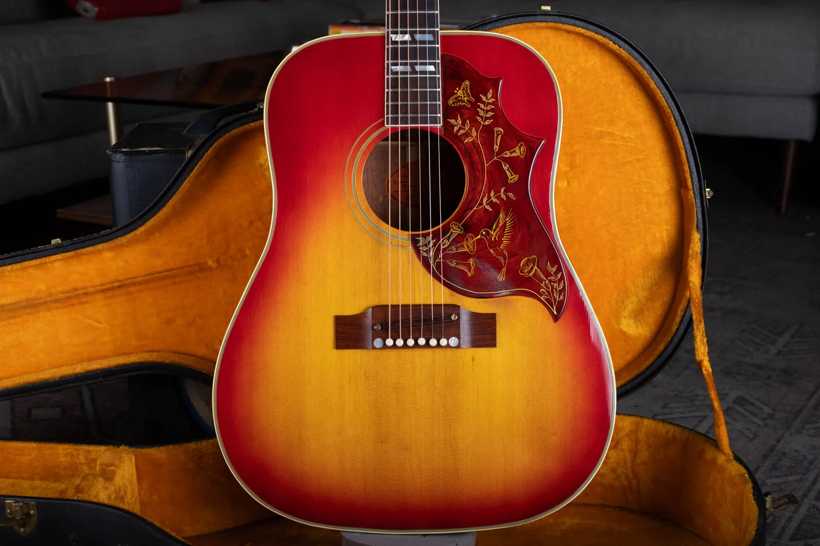 A mid 1960s Gibson Hummingbird in cherry sunburst sitting in its original gold plush lined hardshell case, showing the engraved pickguard and adjustable bridge.