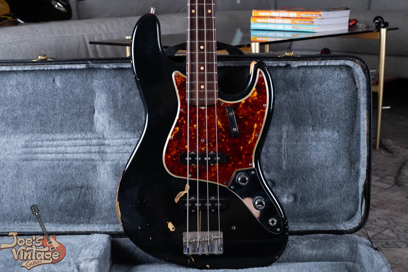 A refinished 1961 Fender Jazz Bass stack knob in black with a tortoiseshell pickguard, standing in a grey lined case, with heavy wear showing bare wood directly beneath the black paint and no undercoat layer