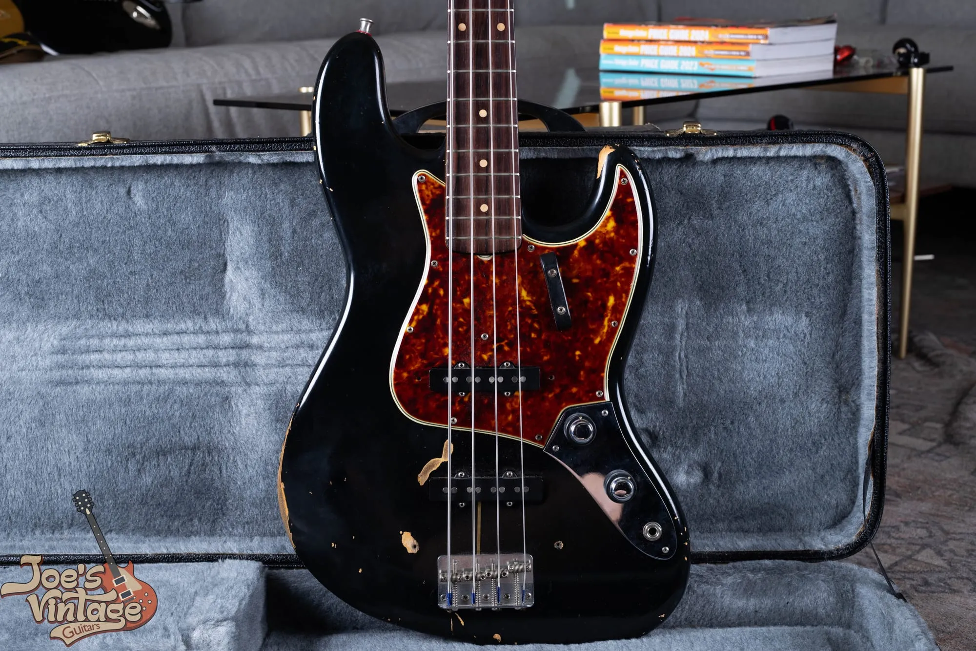A refinished 1961 Fender Jazz Bass stack-knob in black with a tortoiseshell pickguard, standing in a grey-lined case, with heavy wear showing bare wood directly beneath the black paint and no undercoat layer.