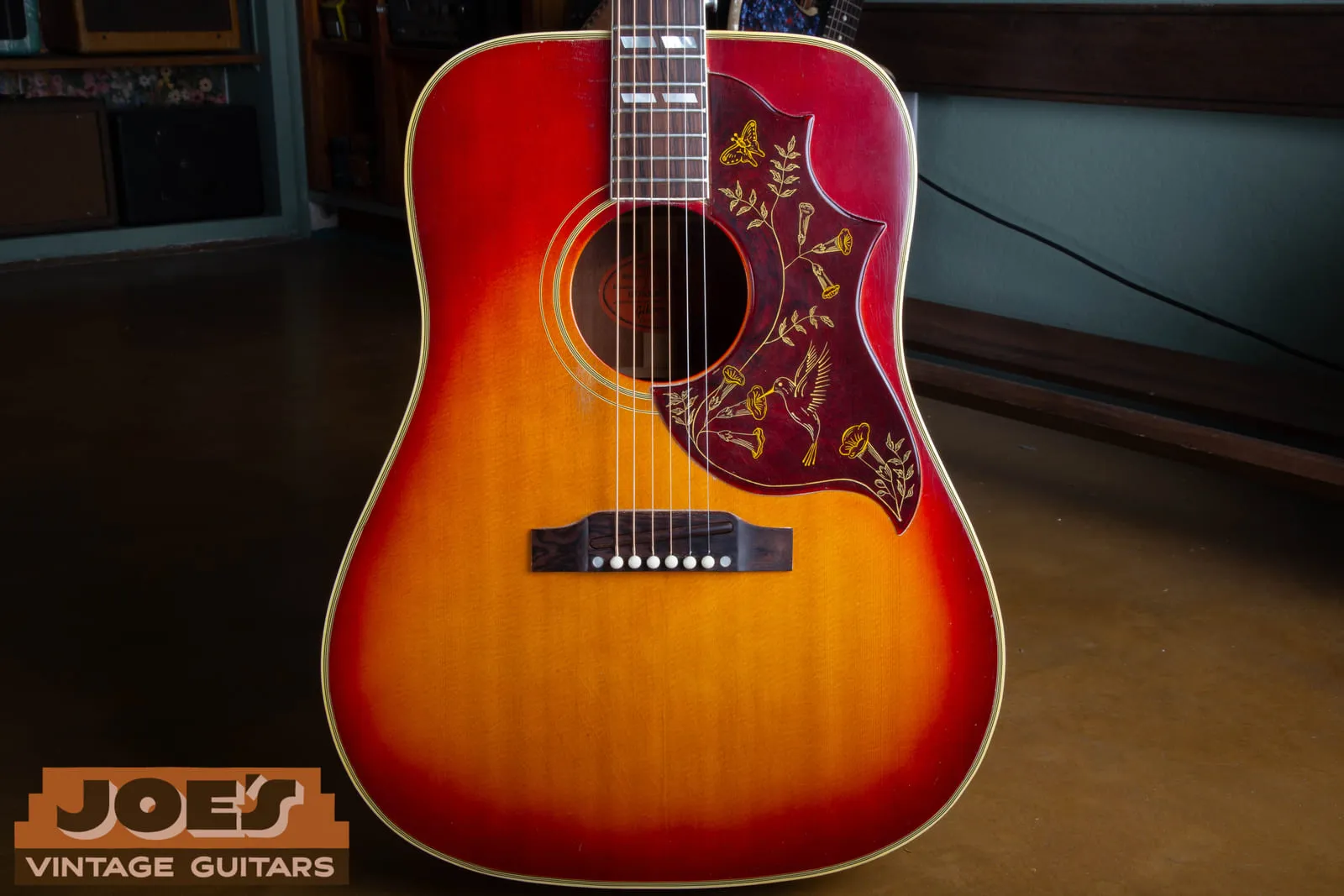 Front view of a 1961 Gibson Hummingbird in cherry sunburst, showing the engraved pickguard with hummingbird, trumpet flowers and butterfly, parallelogram fretboard inlays, and rectangular rosewood bridge.