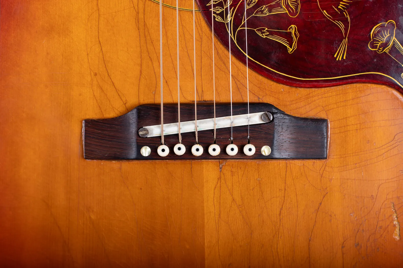 Close up of the adjustable bridge on a 1964 Gibson Hummingbird with a white ceramic saddle sitting on two brass adjustment screws, surrounded by heavy finish checking on the spruce top.