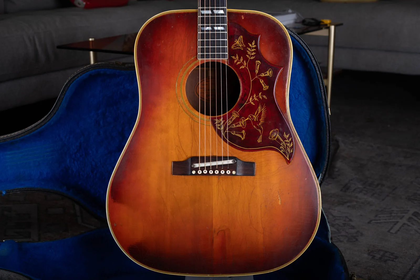 Front of a well played 1964 Gibson Hummingbird with a faded amber sunburst top, heavy finish checking, adjustable bridge, and engraved pickguard, sitting in a blue lined case.