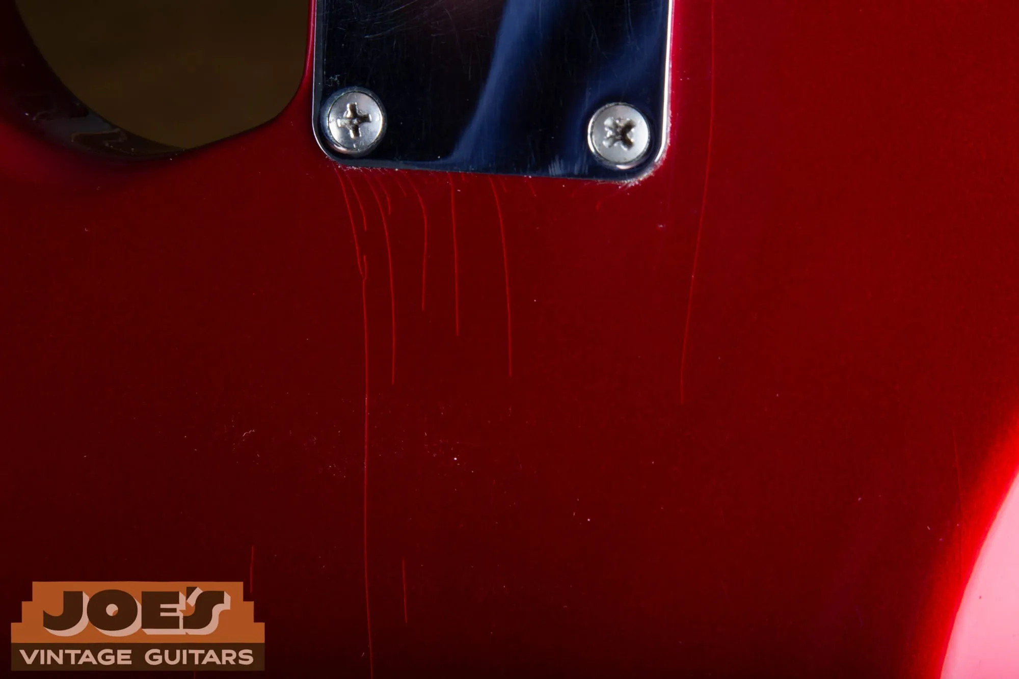 Extreme close-up of Candy Apple Red finish beside the neck plate of a 1965 Fender Stratocaster, showing fine vertical lacquer checking lines running through the deep red metallic finish.
