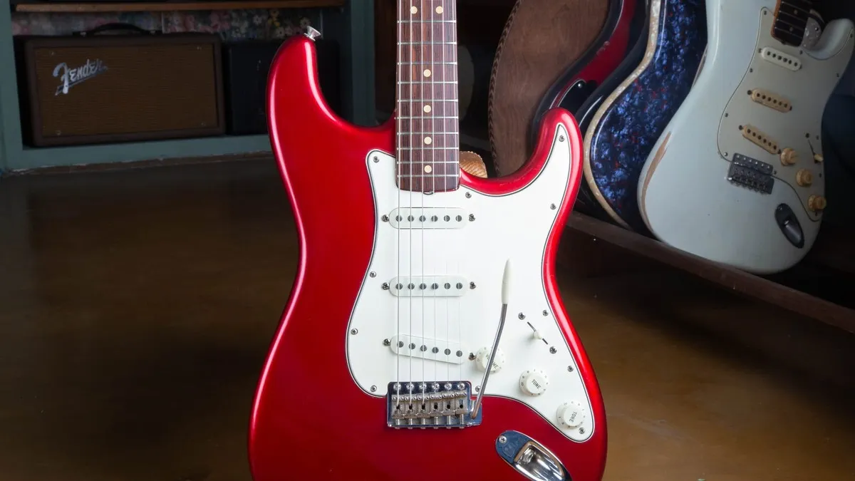 1965 Fender Stratocaster in Candy Apple Red Metallic with a white three-ply pickguard, standing in a shop in front of a blackface Fender amp and other vintage Fenders.