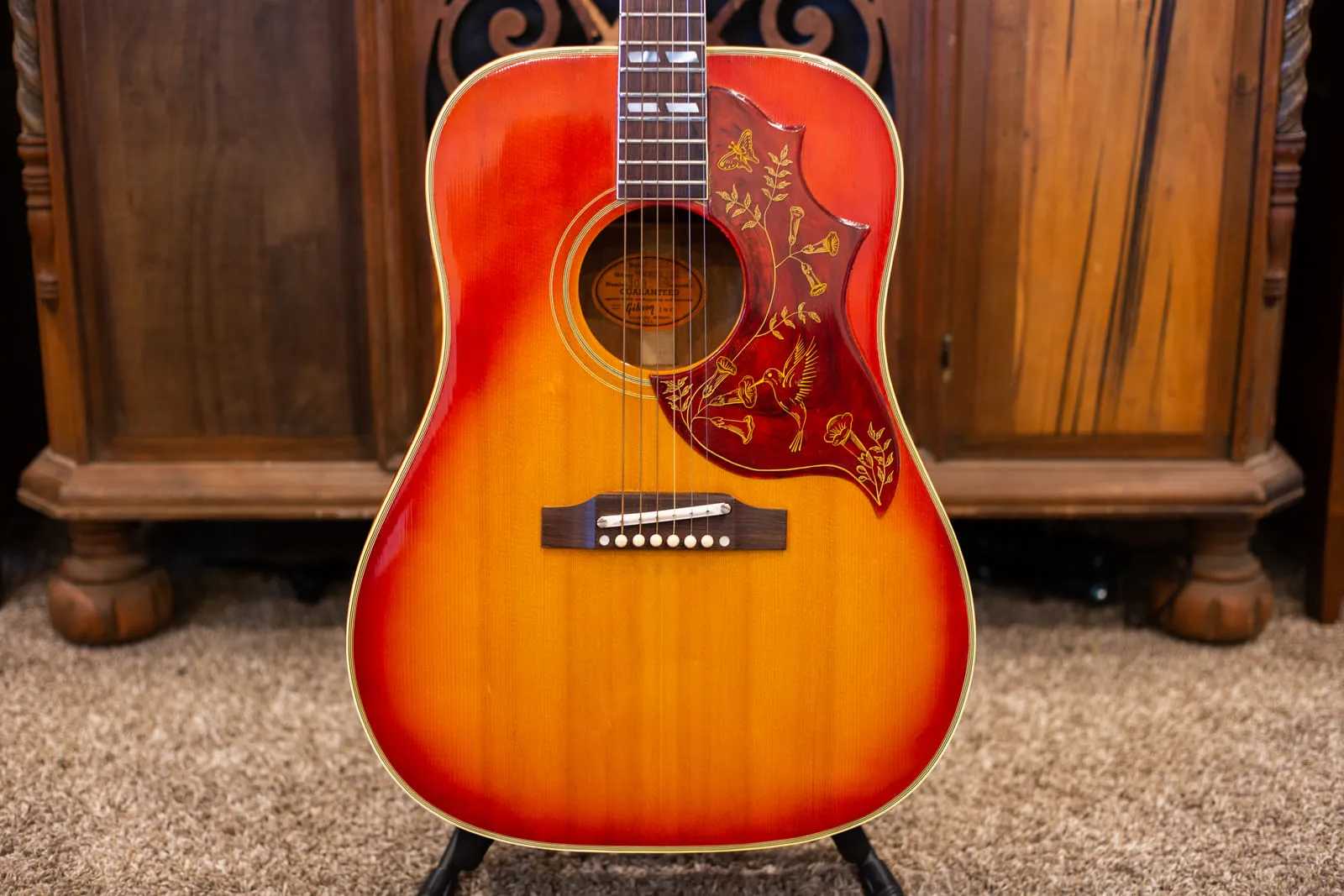 Front view of a 1965 Gibson Hummingbird in cherry sunburst with an adjustable bridge and white ceramic saddle, photographed against a carved wooden cabinet.