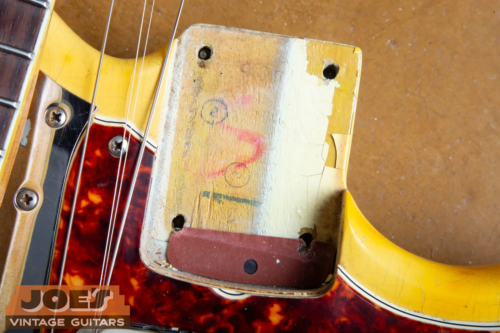 The neck pocket of a 1966 Fender Jaguar in Olympic White showing factory crayon markings, a fiber shim, and a whiter band of paint at one edge where the clear coat has ambered elsewhere