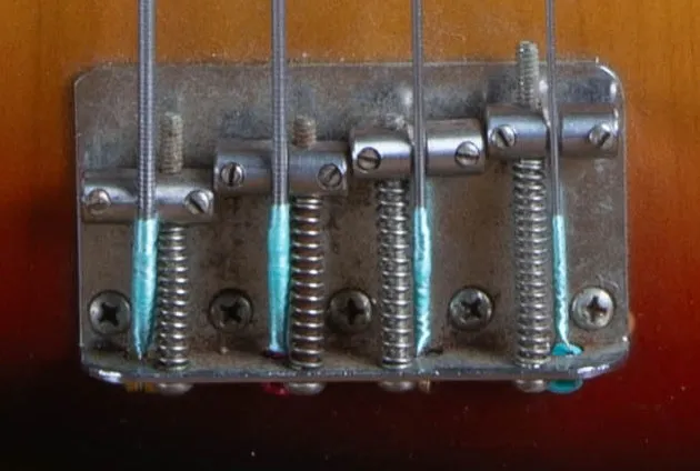 The chrome bridge of a 1973 Fender Jazz Bass with four threaded saddles and the strings anchored through the bridge plate