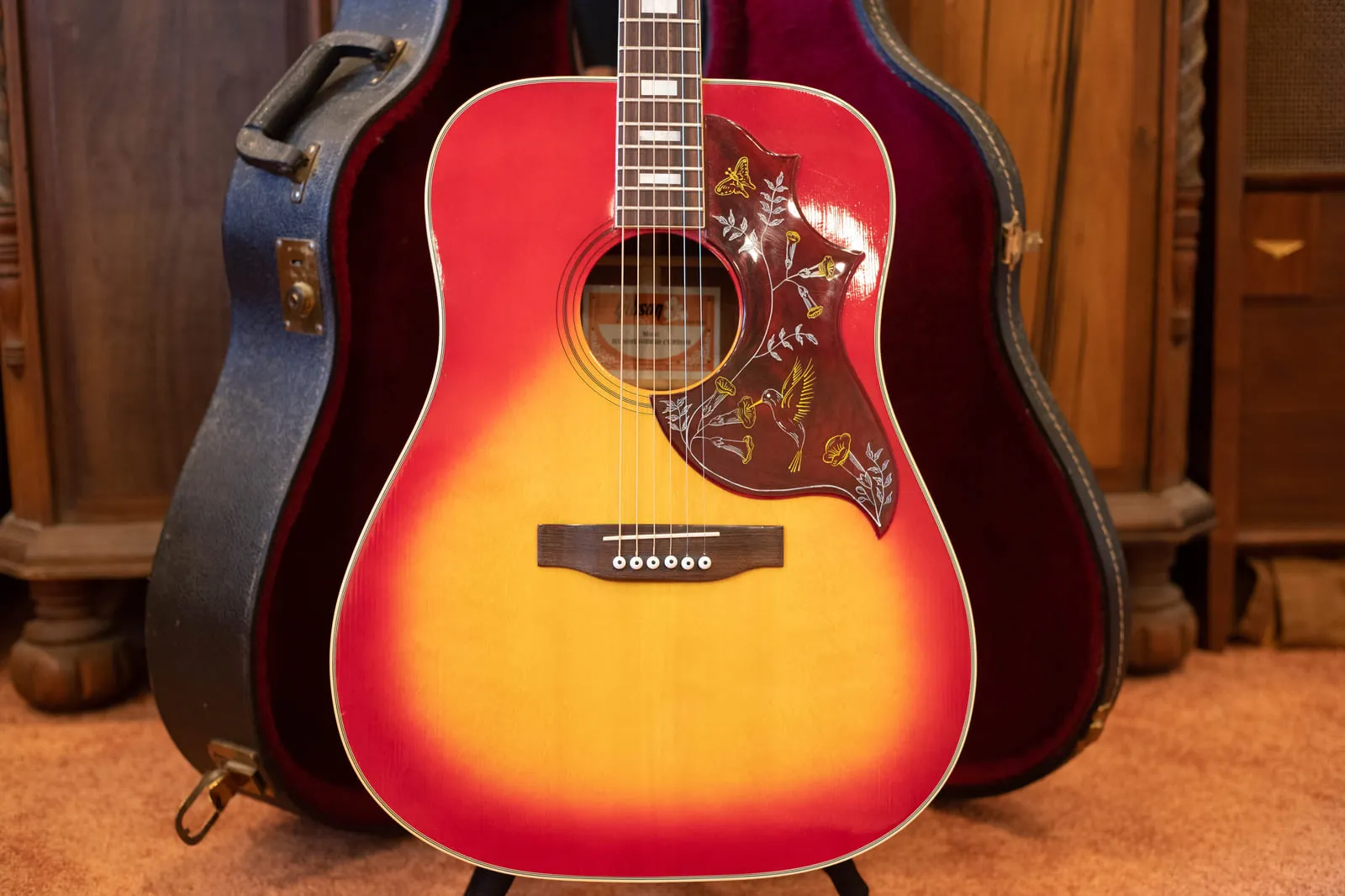 Front of a 1979 Gibson Hummingbird Custom in cherry sunburst with a fixed saddle bridge and a pickguard engraved in white and gold, sitting in front of its blue hardshell case.