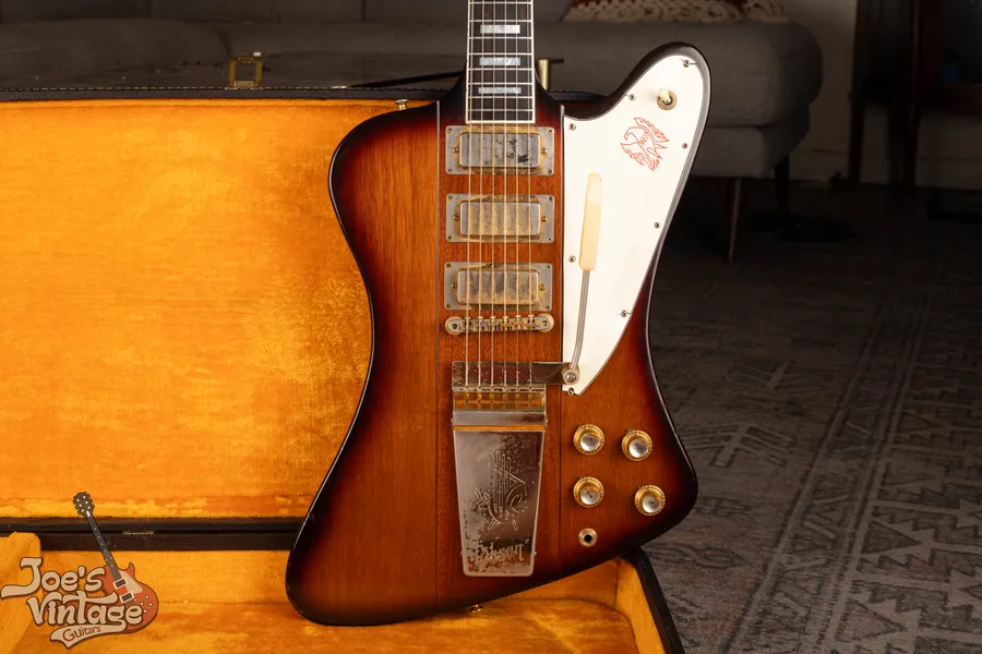 Sunburst Firebird body with three covered pickups and a white pickguard beside an orange-lined case