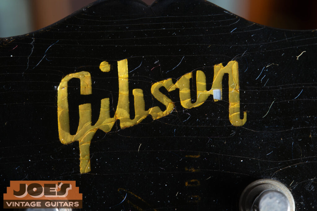 Macro close-up of a 1957 Gibson Les Paul headstock showing a Mother of Pearl "Gibson" logo with significant finish checking above it and a faded gold "Les Paul Model" silkscreen.