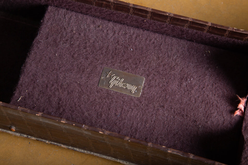 Detailed macro photograph of the original bronze-colored Gibson logo plaque located on the interior neck rest of an alligator chipboard case. The small, rectangular metal plate features the "Gibson" script in relief accompanied by a distinct five-pointed star icon, showing a natural aged patina and minor oxidation consistent with a 1950s accessory.