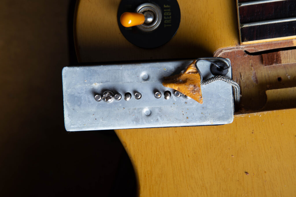 Macro view of the underside of an original 1958 Gibson P-90 soapbar pickup. The image highlights the metal baseplate with the signature little rubber grommet where the lead wires exit, along with the period-correct masking tape used to secure and protect the wiring.