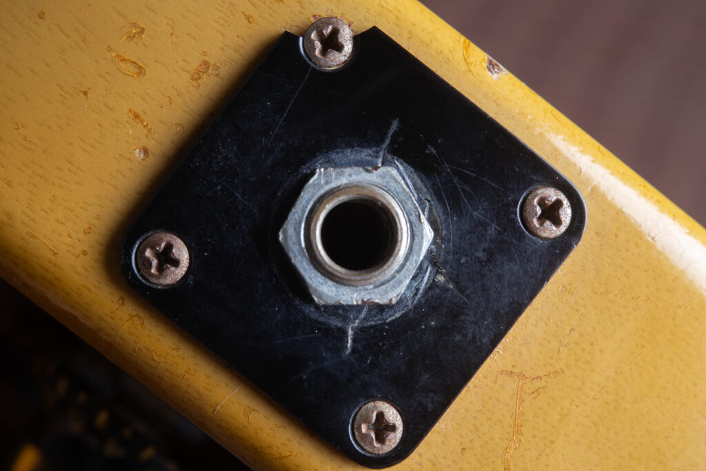 Detailed perspective of the side-mounted output jack on a vintage Gibson Les Paul Special. The image focuses on the original black plastic square jack plate, secured by four nickel-plated screws. The surrounding TV Yellow "limed mahogany" finish shows authentic nitrocellulose weather checking and natural patina around the plate edges.